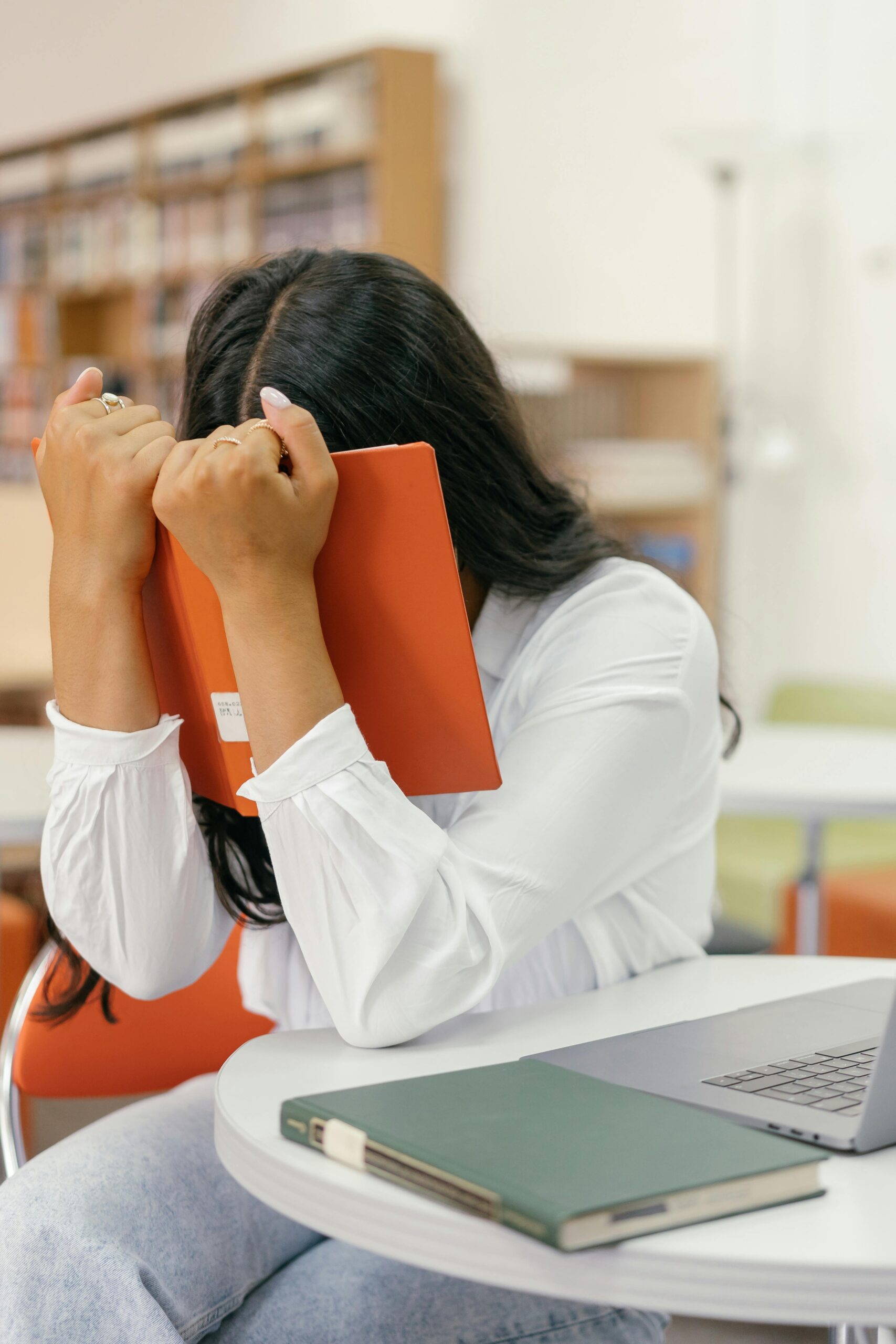 Young woman in library holds head in frustration behind book, depicting study stress.